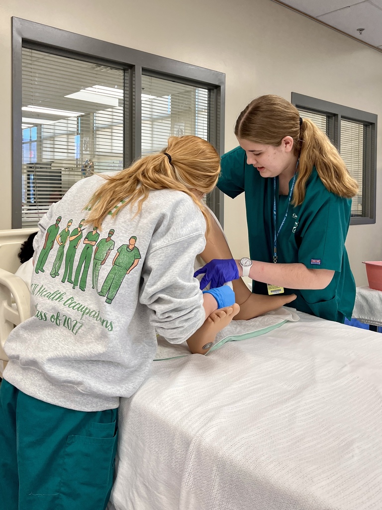 Two students in gloves perform catheter care on a mannequin in a simulated hospital setting.