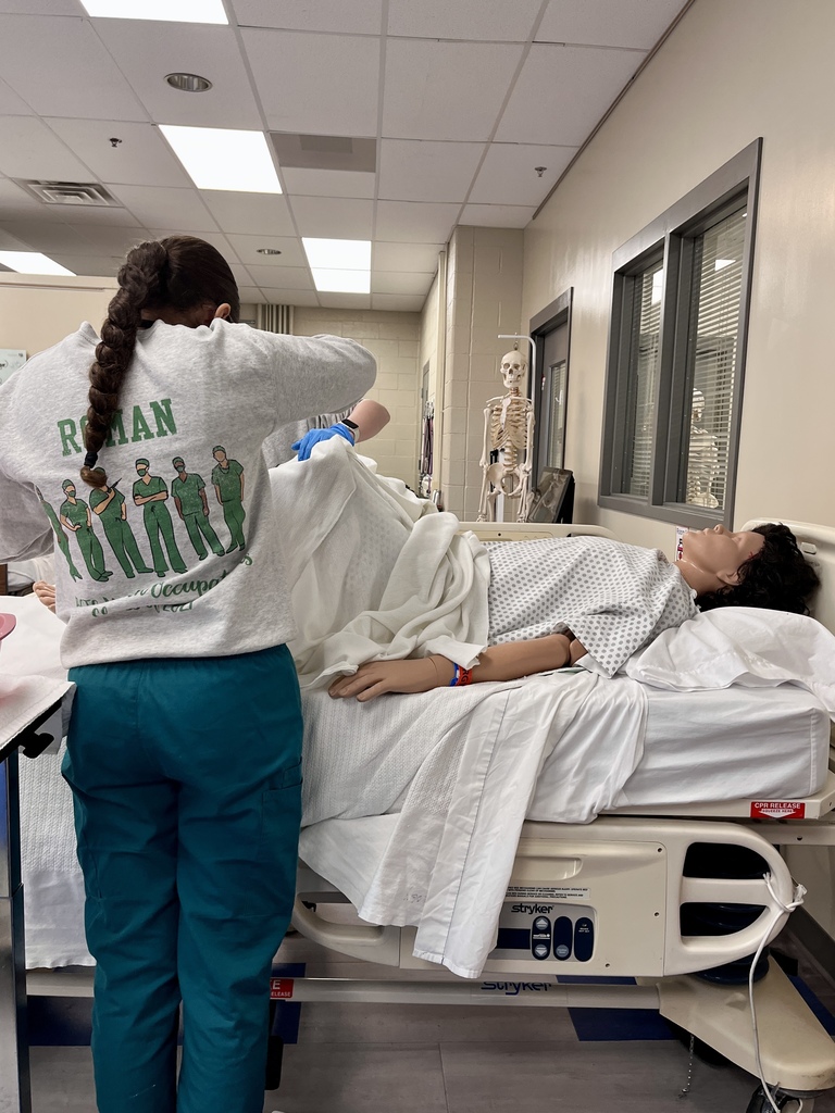 Student adjusts linens on a hospital bed with a mannequin patient in a healthcare training lab.
