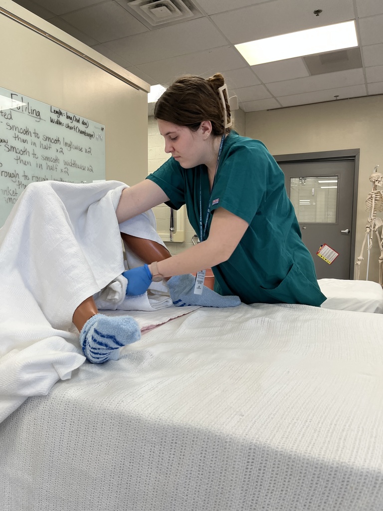 Student in scrubs provides catheter care on a mannequin under a blanket in a healthcare training lab.
