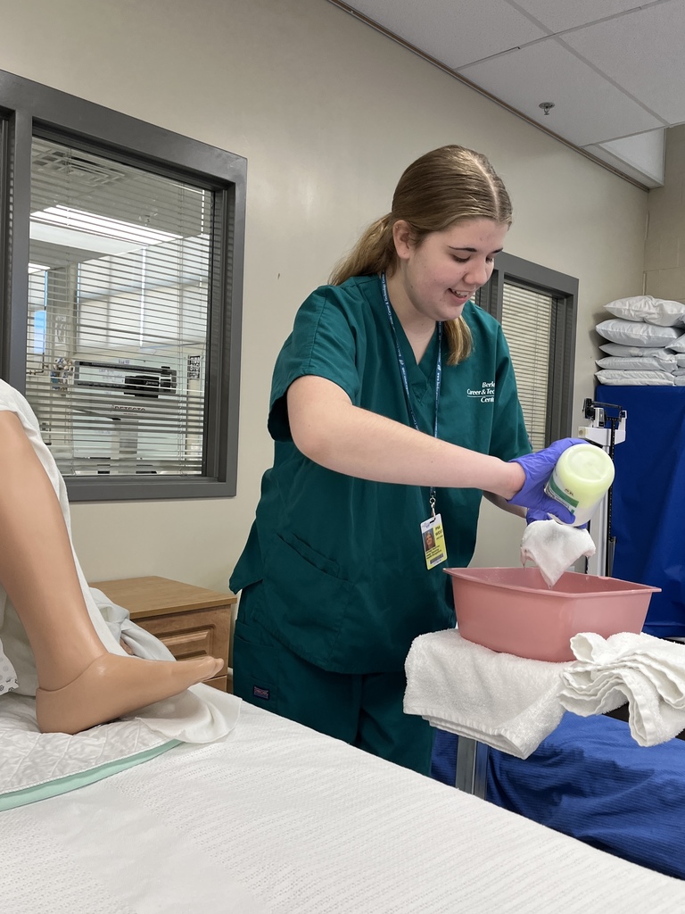 Student in scrubs prepares washcloths with soap at a bedside for patient care practice.