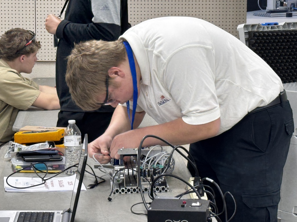Student in safety glasses works on wiring components at a competition table with tools and equipment nearby.