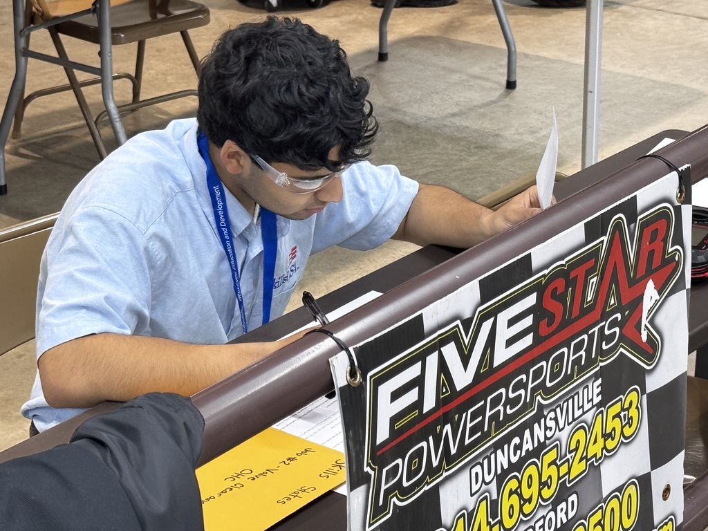 Student wearing safety glasses reviews paperwork at a competition station with a Five Star Powersports sign in front.