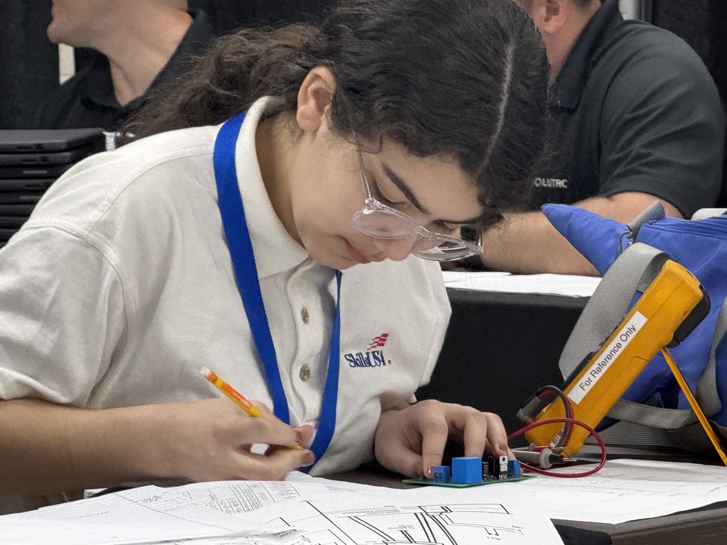 Student in safety glasses carefully works on a small circuit board while referencing a technical drawing.