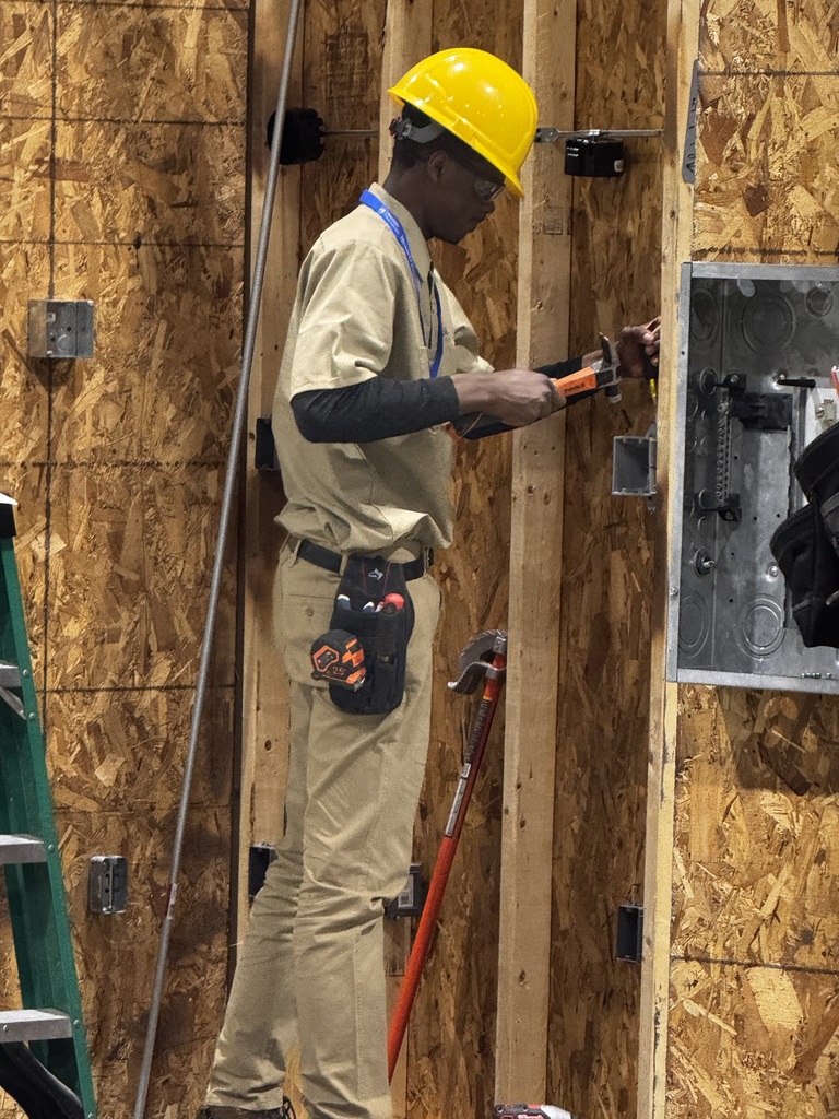 Student wearing a hard hat uses tools to install electrical components inside a framed wall structure.