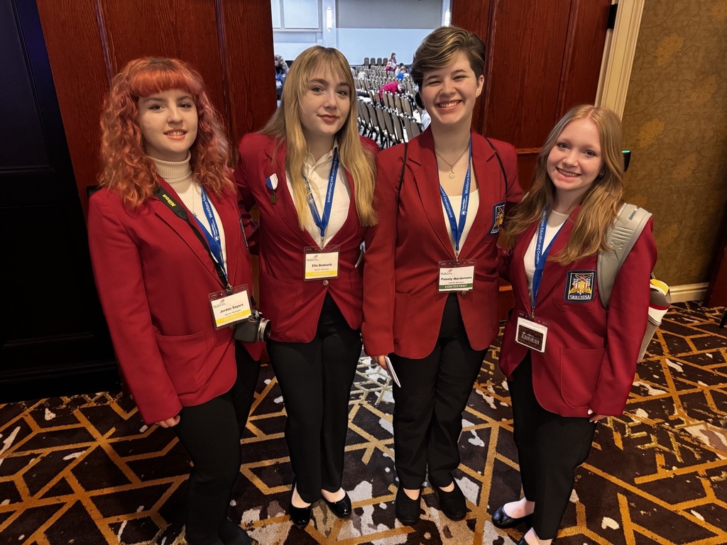 Four SkillsUSA students in red blazers stand together smiling in a conference hallway with seating visible behind them.