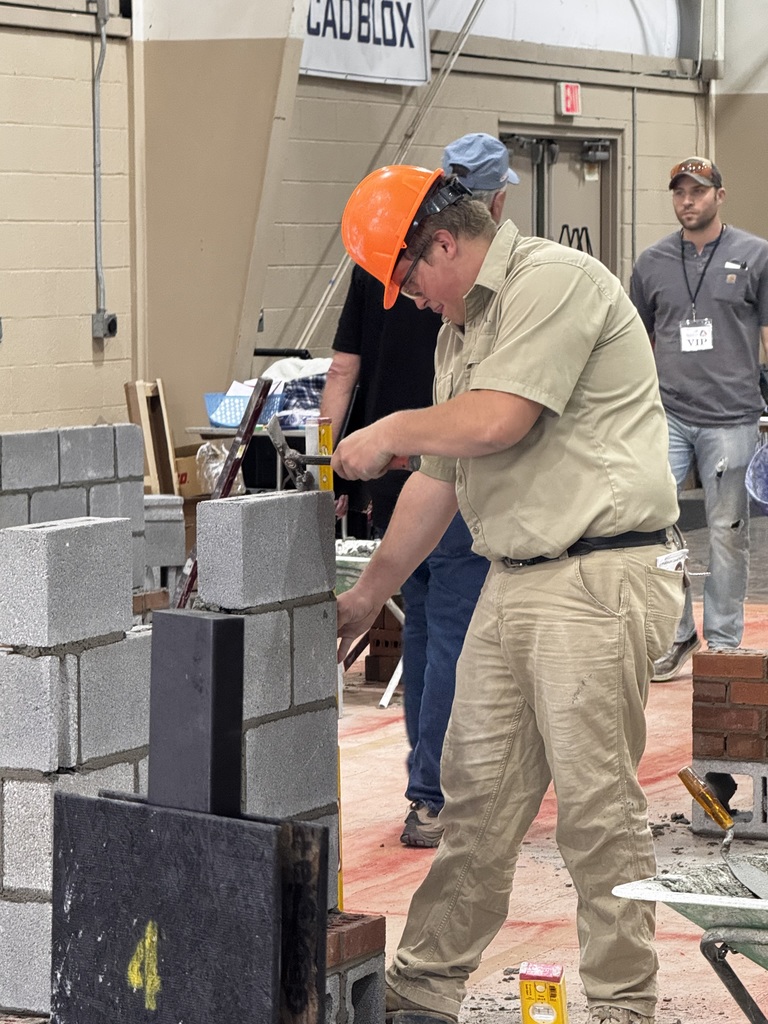 Student in a hard hat builds a concrete block structure during a masonry competition while others observe nearby.