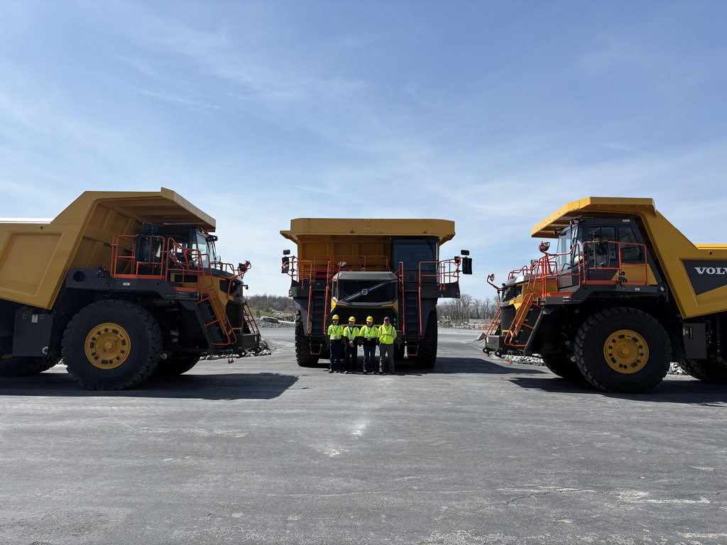Group of individuals in safety gear stand between two large haul trucks at an outdoor quarry site under a clear sky.