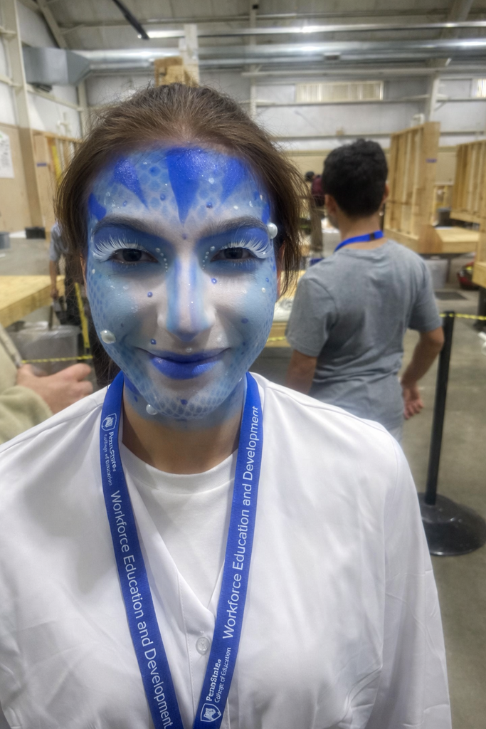 Student wearing detailed blue-and-white fantasy face paint and a white draped costume smiles at the camera inside a large workshop or competition space, with construction framing and other participants visible in the background.