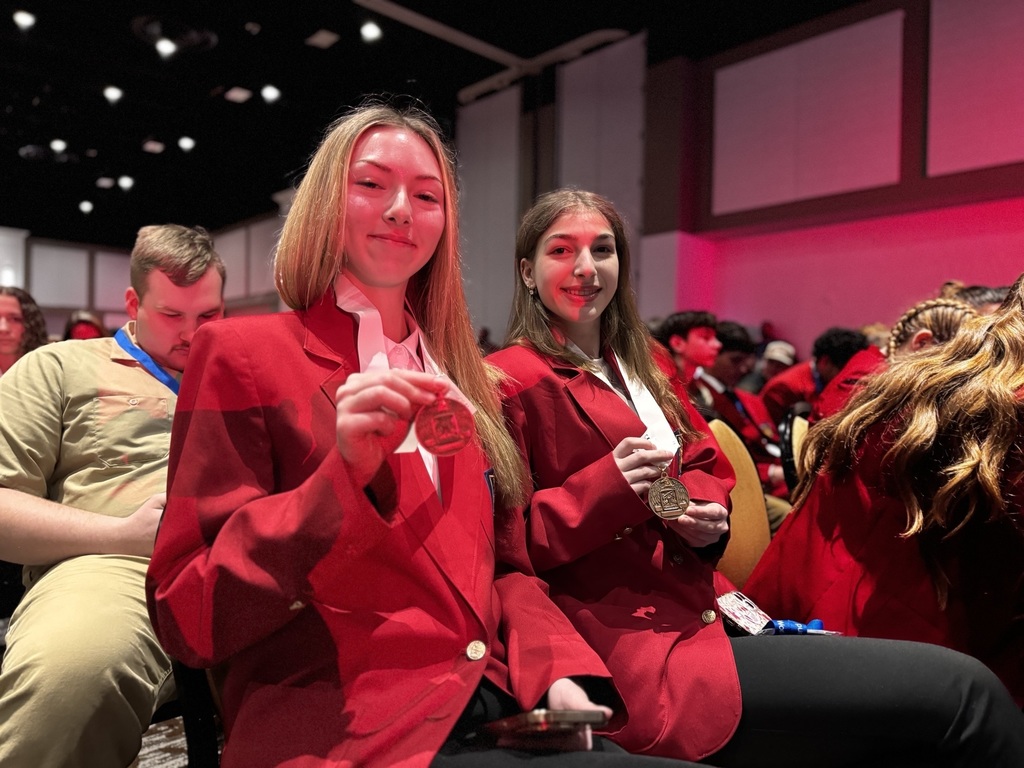 Two BCTC students in red SkillsUSA blazers sit together at the ceremony holding up their medals and smiling for the camera.
