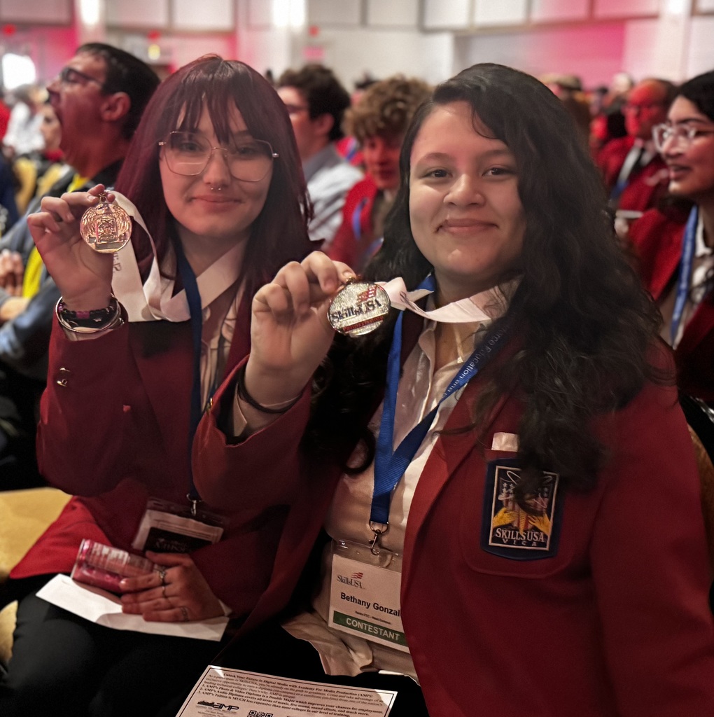 Two BCTC students in red SkillsUSA blazers sit in the audience at the awards ceremony, smiling and holding up their medals while surrounded by other student competitors.