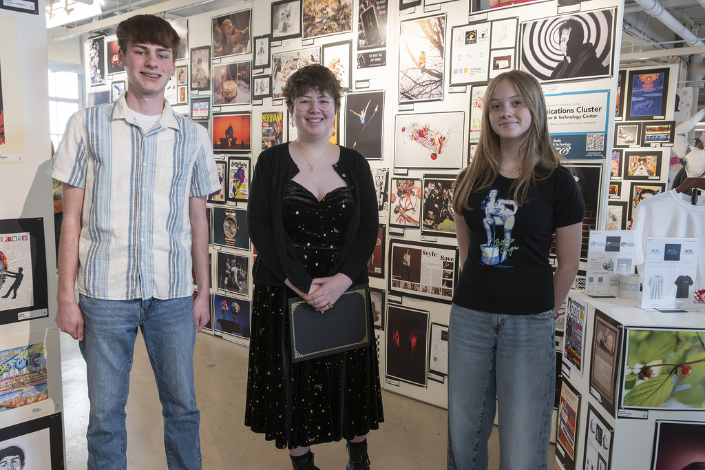 Three students stand in front of a gallery wall filled with student photography and graphic design work at an art exhibit.
