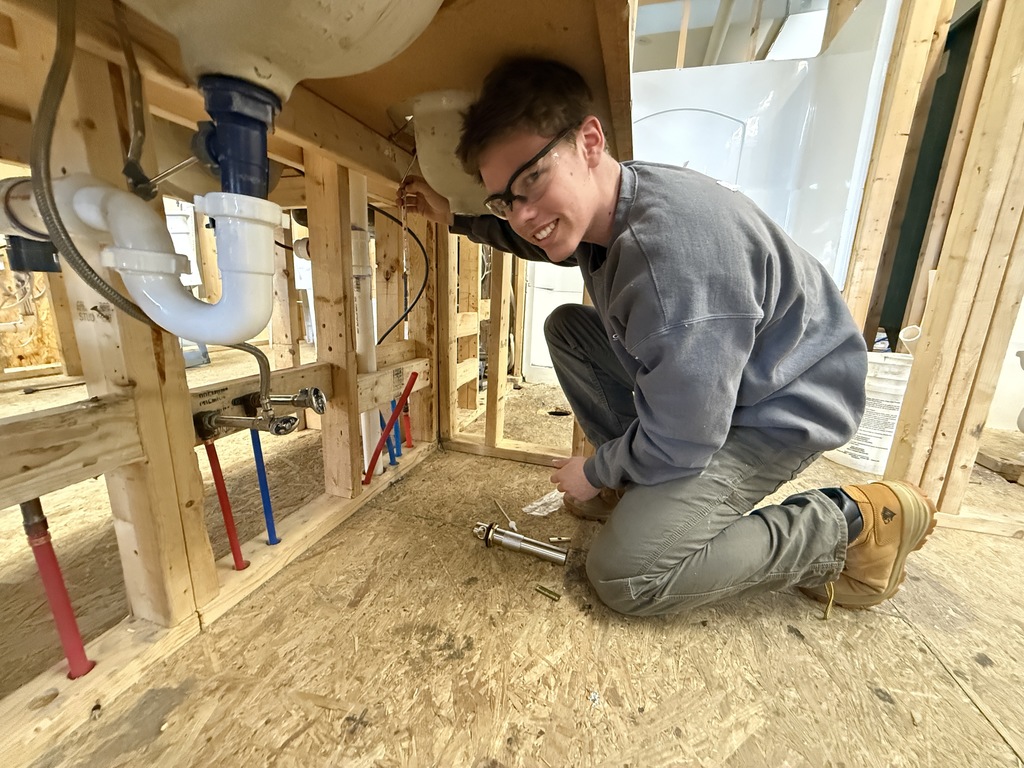 Student working under a sink inside a framed structure, connecting drain and water supply lines.