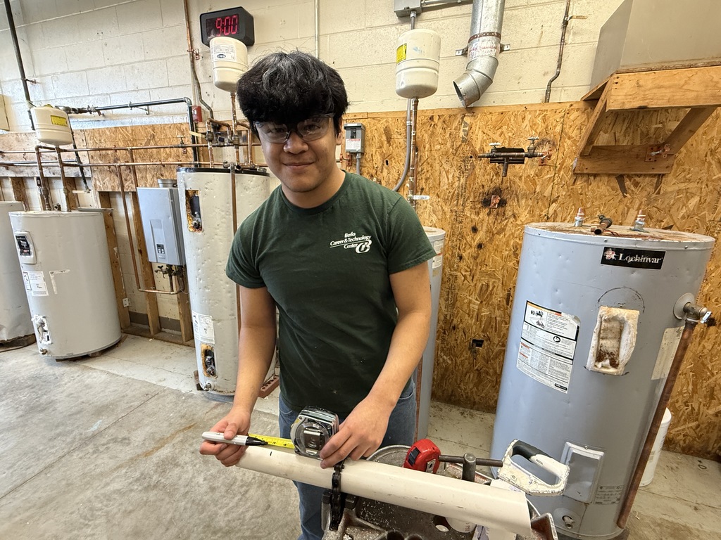 Student in a workshop measures a section of pipe near water heaters and plumbing equipment.