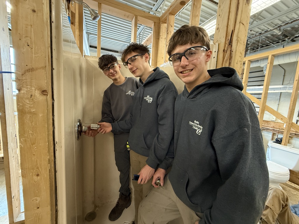 Three students wearing safety glasses stand in a framed shower area, installing plumbing fixtures on the wall.