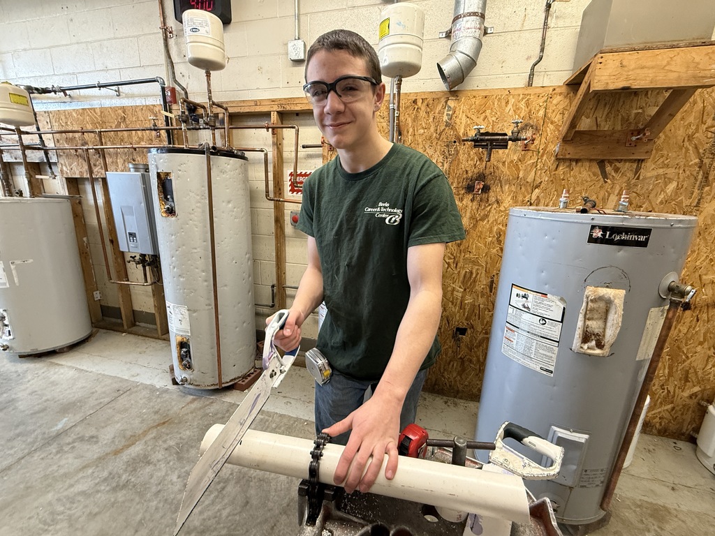Student uses a hand saw to cut a section of pipe while holding it steady on a workbench.
