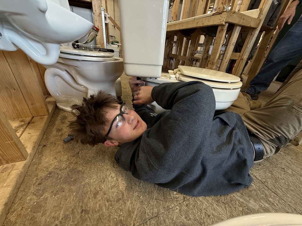 Student lies on the floor connecting plumbing to the base of a toilet during installation.