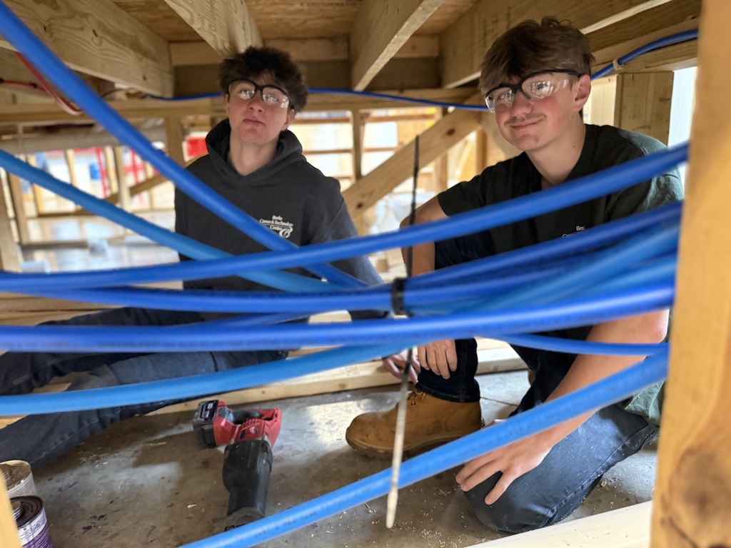 Two students sit beneath a framed structure, working with blue PEX water lines and a power tool nearby.
