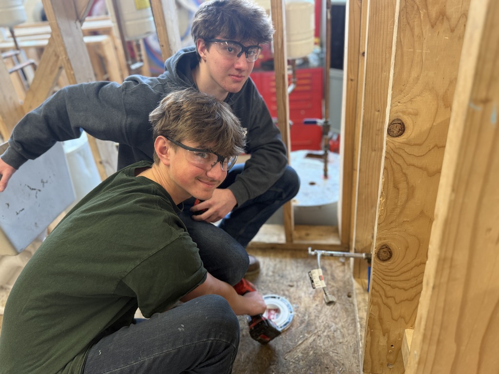 Two students wearing safety glasses kneel beside a framed wall, working on plumbing connections near a floor drain.