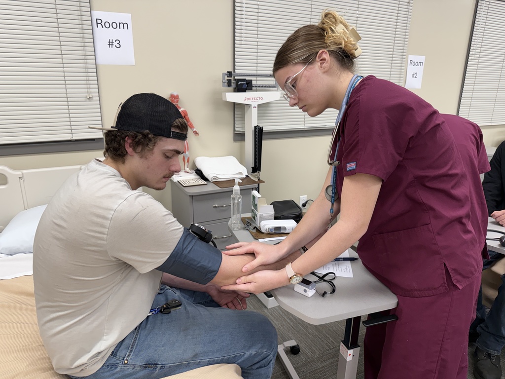 Student in maroon scrubs checks a classmate’s blood pressure while supporting their arm at a bedside station.