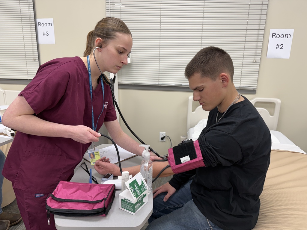 Student in maroon scrubs uses a stethoscope and blood pressure cuff to check a classmate’s vitals in a healthcare lab setting.