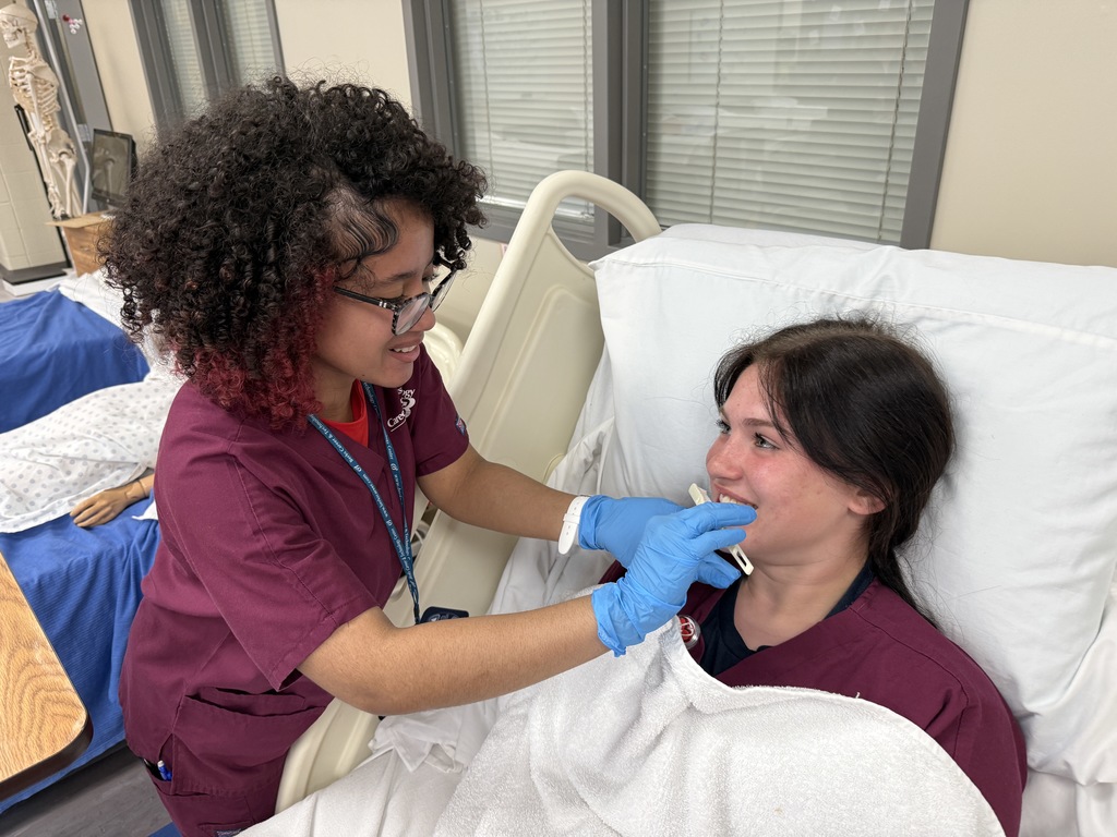 Student wearing gloves assists a classmate with oral care while the patient sits up in a hospital bed.