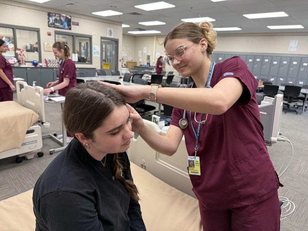 Student in maroon scrubs examines a classmate’s head and face as part of a patient assessment in a classroom lab.