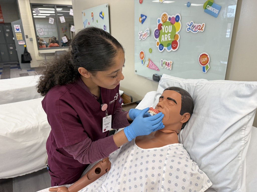Student wearing gloves demonstrates denture cleaning on a medical mannequin in a healthcare classroom.