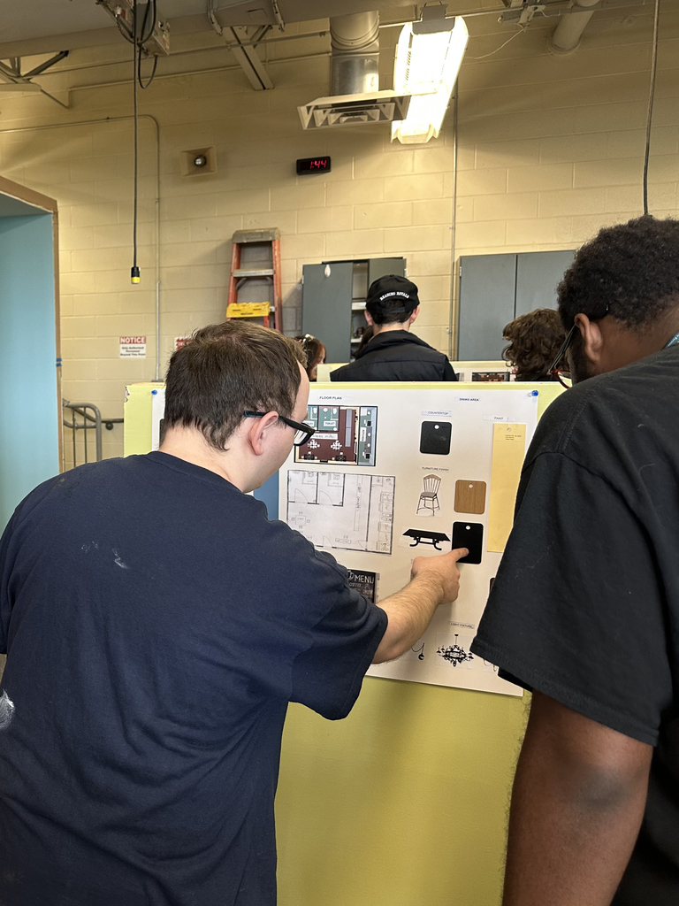 Two students closely examining a design board with floor plans and material samples, pointing and discussing details.