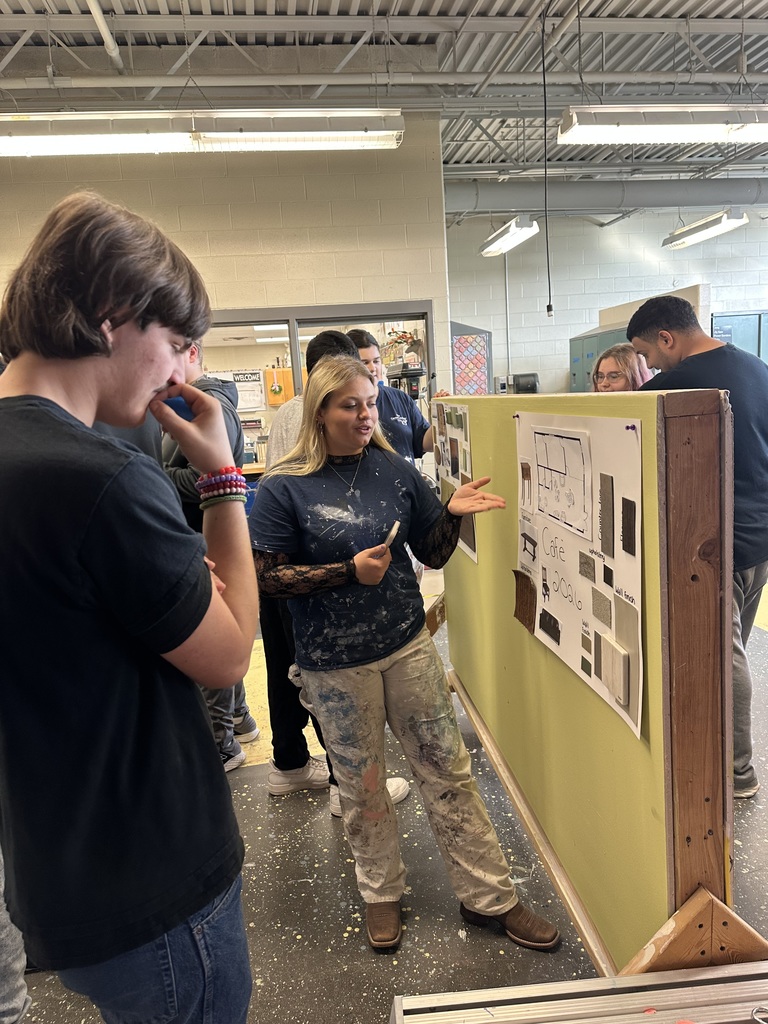 Student presenting a coffee shop design board with layout and material samples while classmates observe and listen.