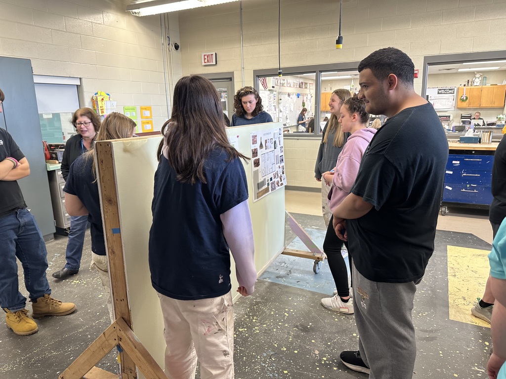 Students gathered around a presentation board reviewing a coffee shop design while listening and discussing ideas in a classroom.