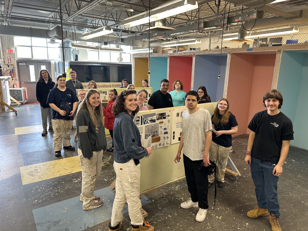 Group of students in a classroom workshop standing around display boards with coffee shop design plans, smiling and posing together.