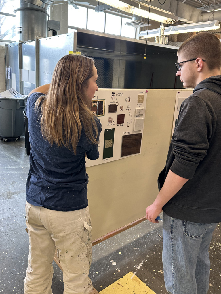 Student explaining a design board with color and material samples to another student during a classroom critique session.