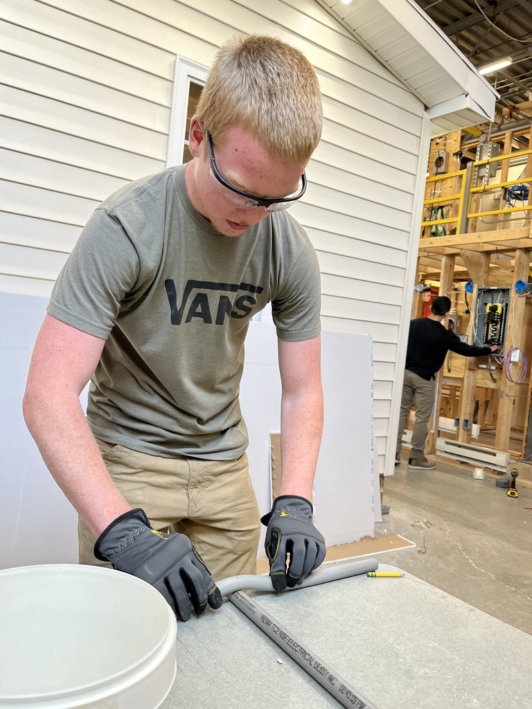 Student bends conduit while another secures it along a framed wall section.