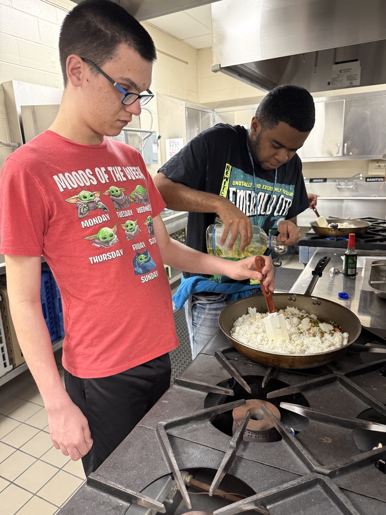 Two students prepare fried rice, adding oil and mixing rice and vegetables in a skillet.