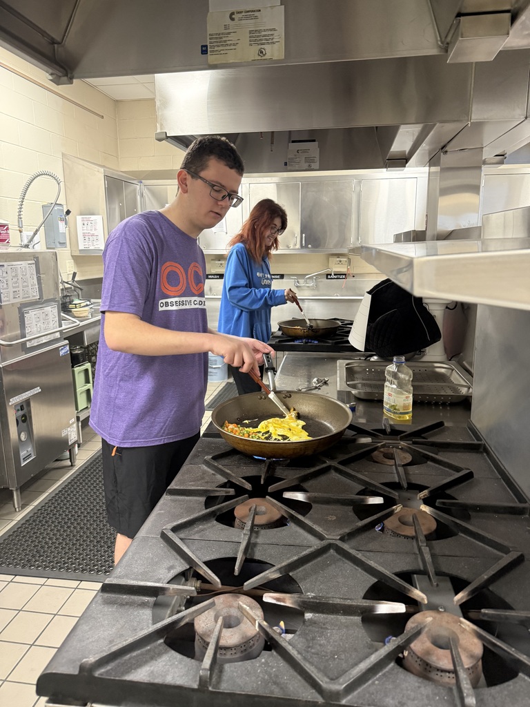 Student stirs eggs and vegetables in a skillet while another student cooks in the background in a commercial kitchen.