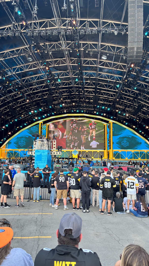 Wide view of the NFL Draft stage with large screens and lighting rig as a crowd of fans watches from behind a barrier.