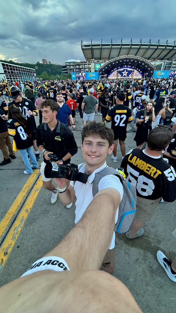 Young man takes a selfie in a large crowd at an outdoor NFL Draft stage, holding a camera, with fans in team jerseys behind him.