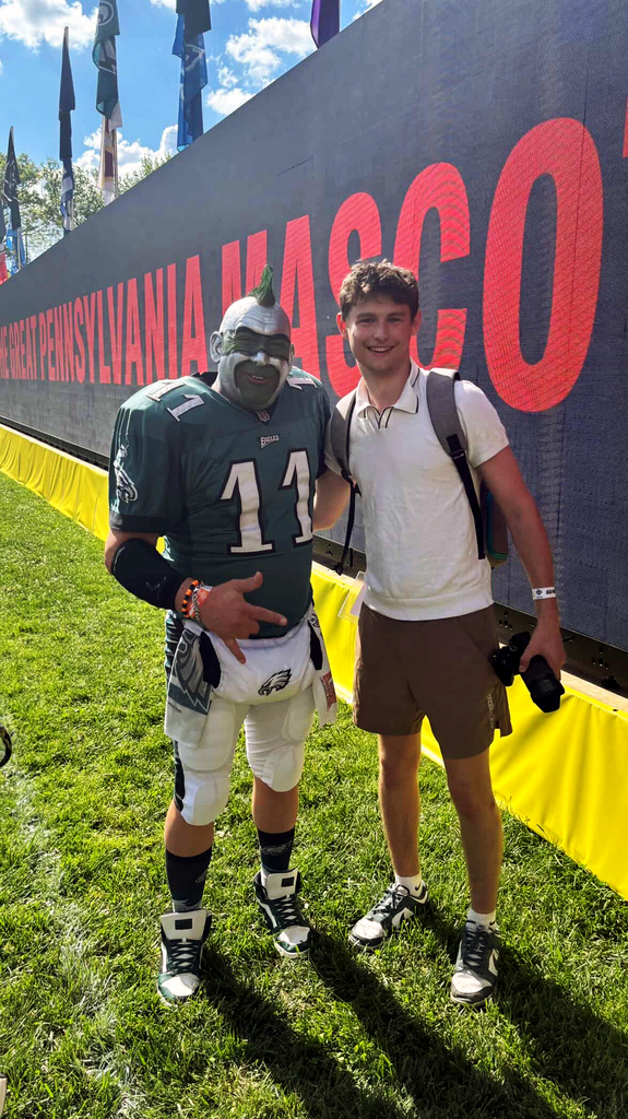 Young man with camera poses next to a Philadelphia Eagles fan in full costume in front of a large event sign.