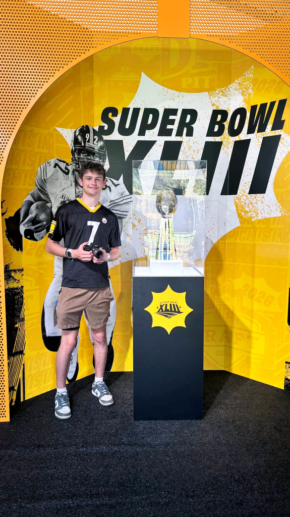 Young man holding a camera stands beside a Super Bowl display case featuring the Lombardi Trophy against a yellow backdrop