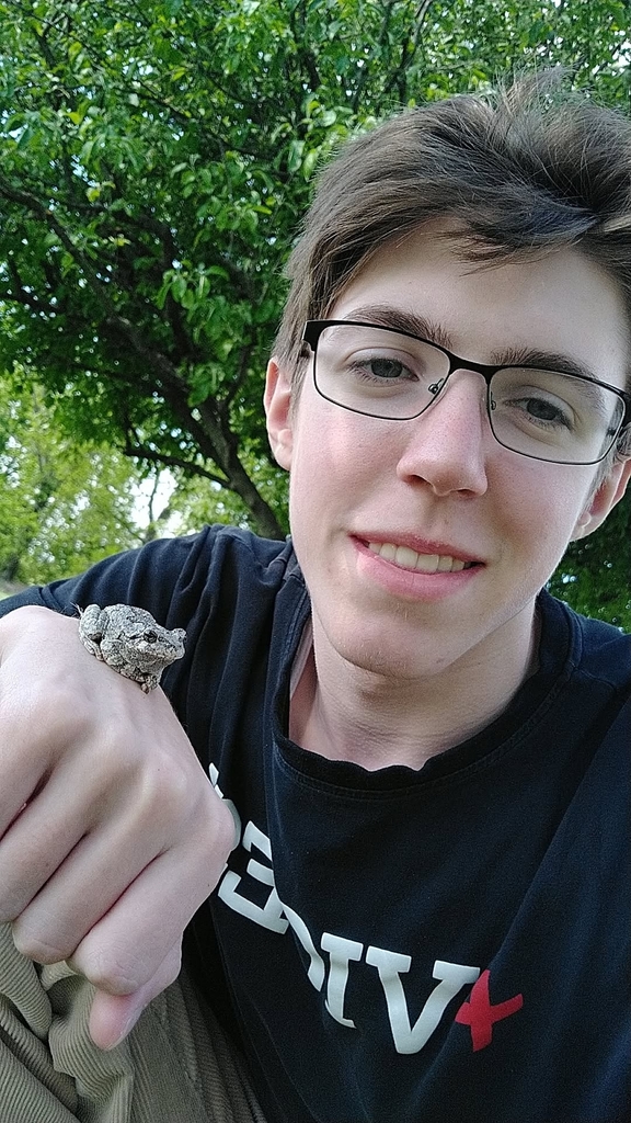 Student wearing glasses smiles outdoors while holding a small gray tree frog on their hand, with leafy green trees in the background.