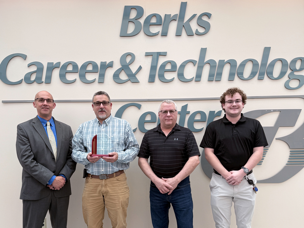 Four men stand in front of a Berks Career & Technology Center wall sign; one holds a red award plaque while the others stand beside him.