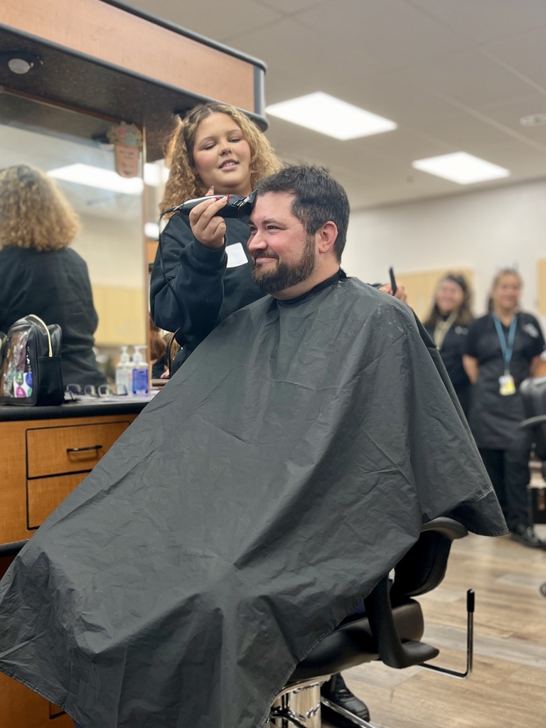 Cosmetology student uses clippers to begin shaving Mr. Entrekin’s hair as classmates watch in the salon.