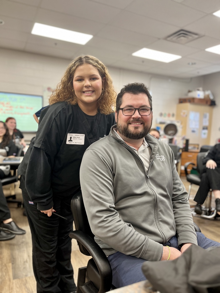Cosmetology student stands beside Mr. Entrekin as he sits in a salon chair before students begin shaving his head.
