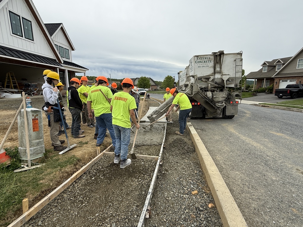 Masonry students in hard hats use rakes and tools to spread freshly poured concrete beside the Student Built House.