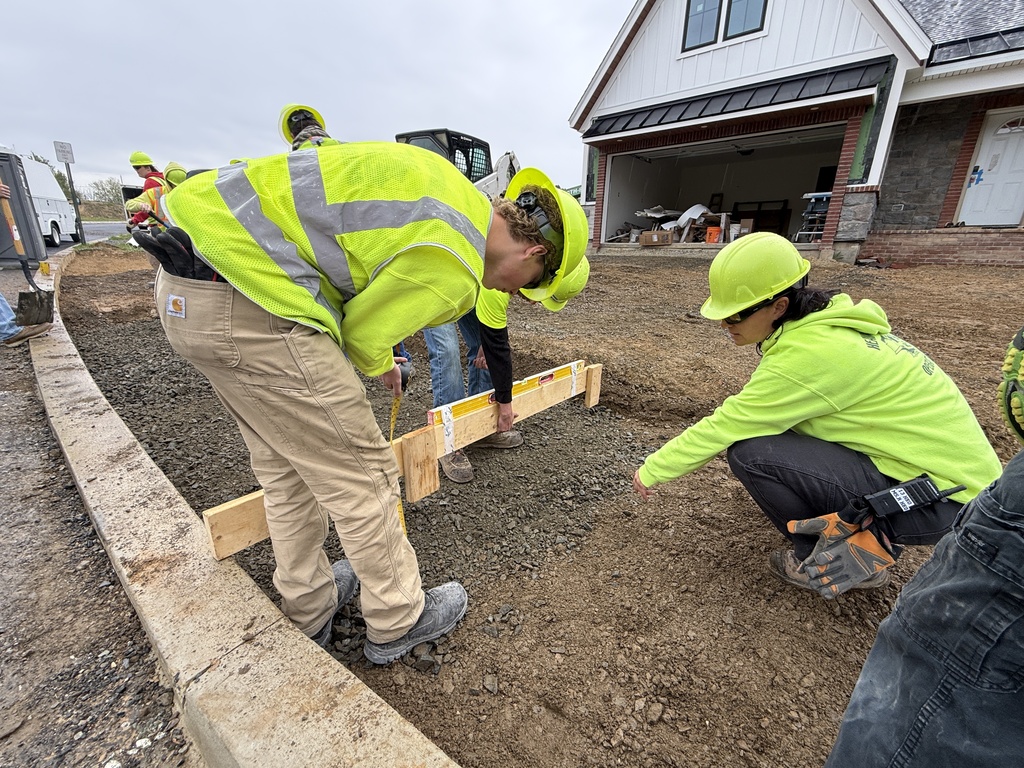 Heavy Equipment Operations students in hard hats and safety vests check and level a gravel base outside the Student Built House.
