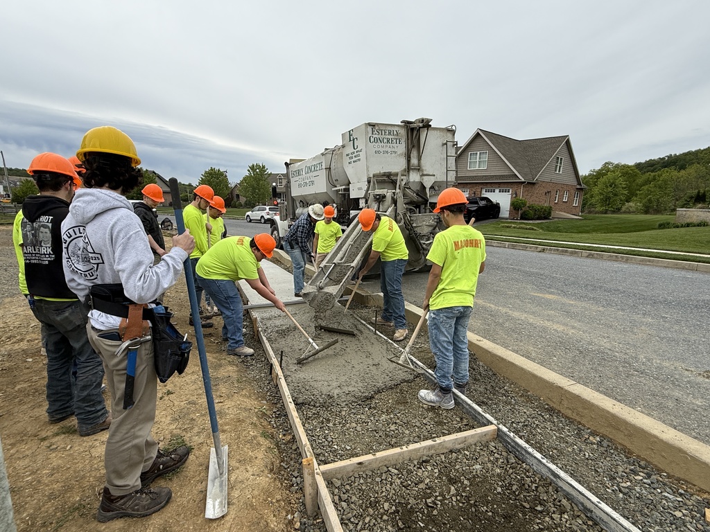 Masonry students level and spread wet concrete as it flows from a concrete truck chute into a framed walkway.