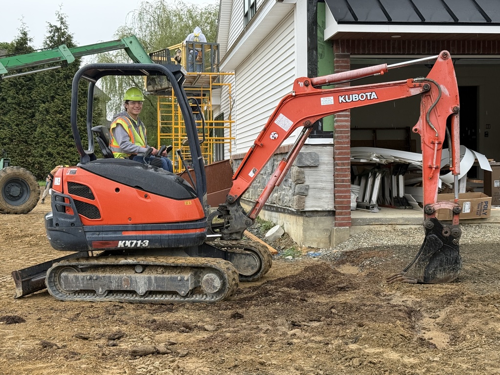 A Heavy Equipment Operations student operates a compact excavator near the Student Built House while classmates work nearby.