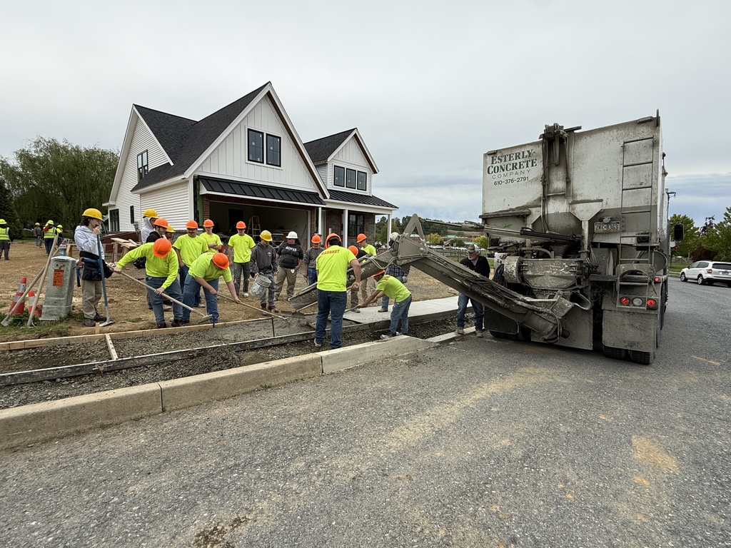 Masonry students work together as a concrete truck pours concrete into a framed walkway near the Student Built House.