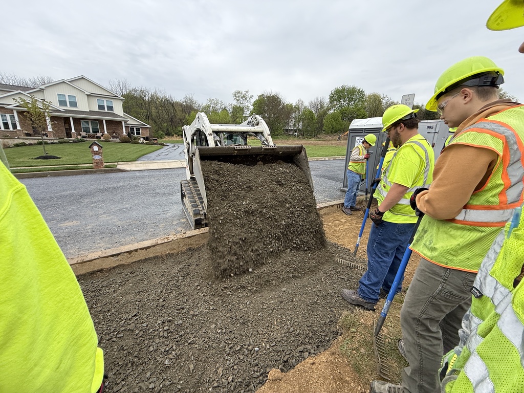 Students in hard hats and safety vests watch as a skid steer dumps gravel into the prepared area near the Student Built House.