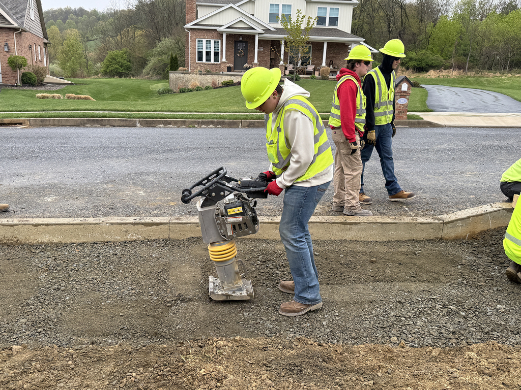 A Heavy Equipment Operations student uses a jumping jack tamper to compact gravel as classmates observe near the Student Built House.